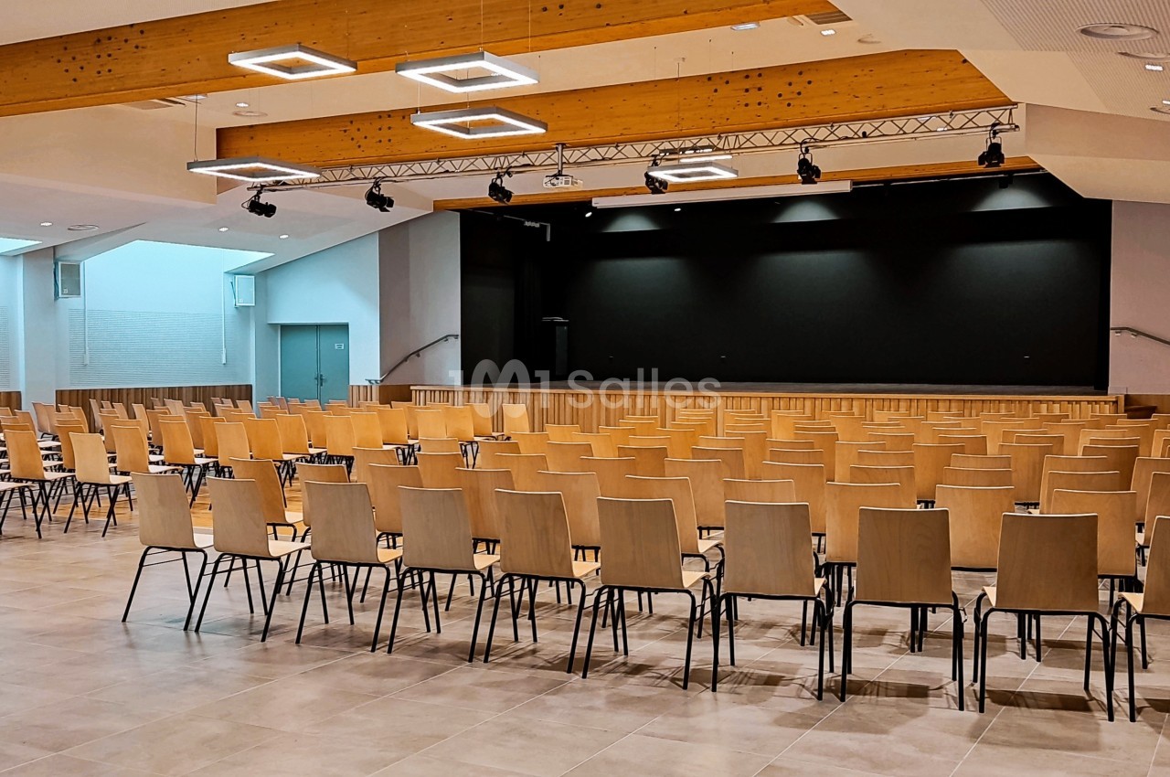 Salle vide avec des rangées de chaises en bois face à une scène noire, éclairée par des plafonniers modernes.