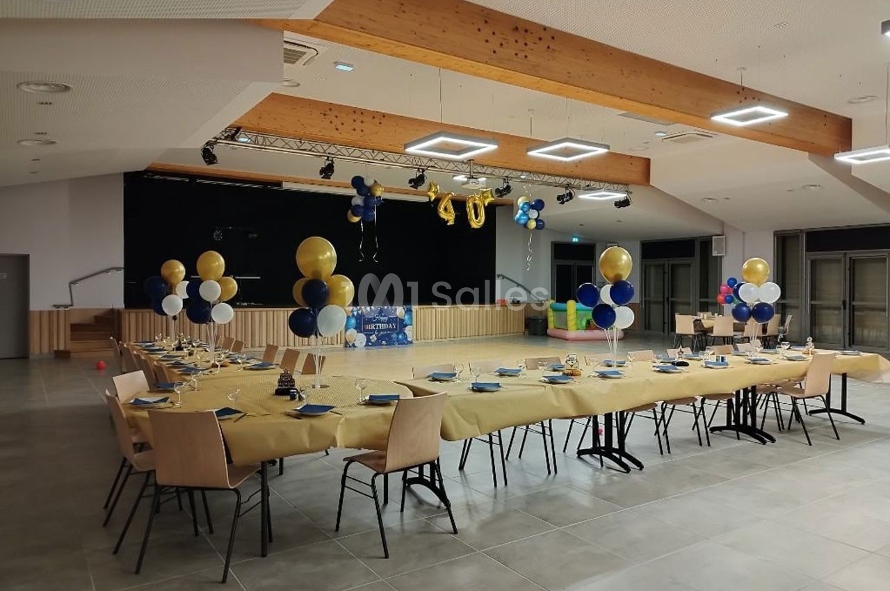 Salle décorée pour un événement avec tables dressées, nappes jaunes, ballons bleus et dorés, et une scène en arrière-plan.