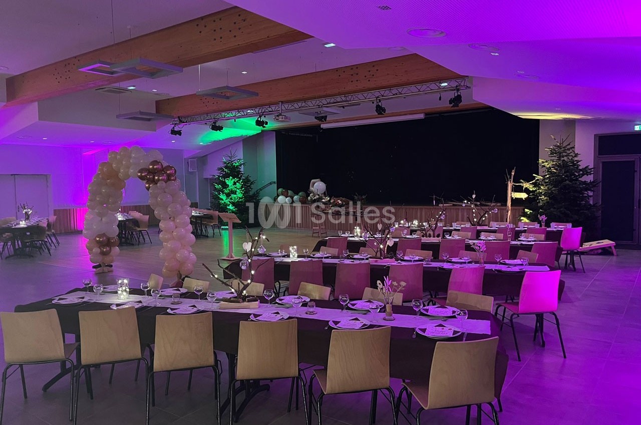 Salle de réception décorée avec des tables dressées, des ballons et un éclairage coloré violet et vert.