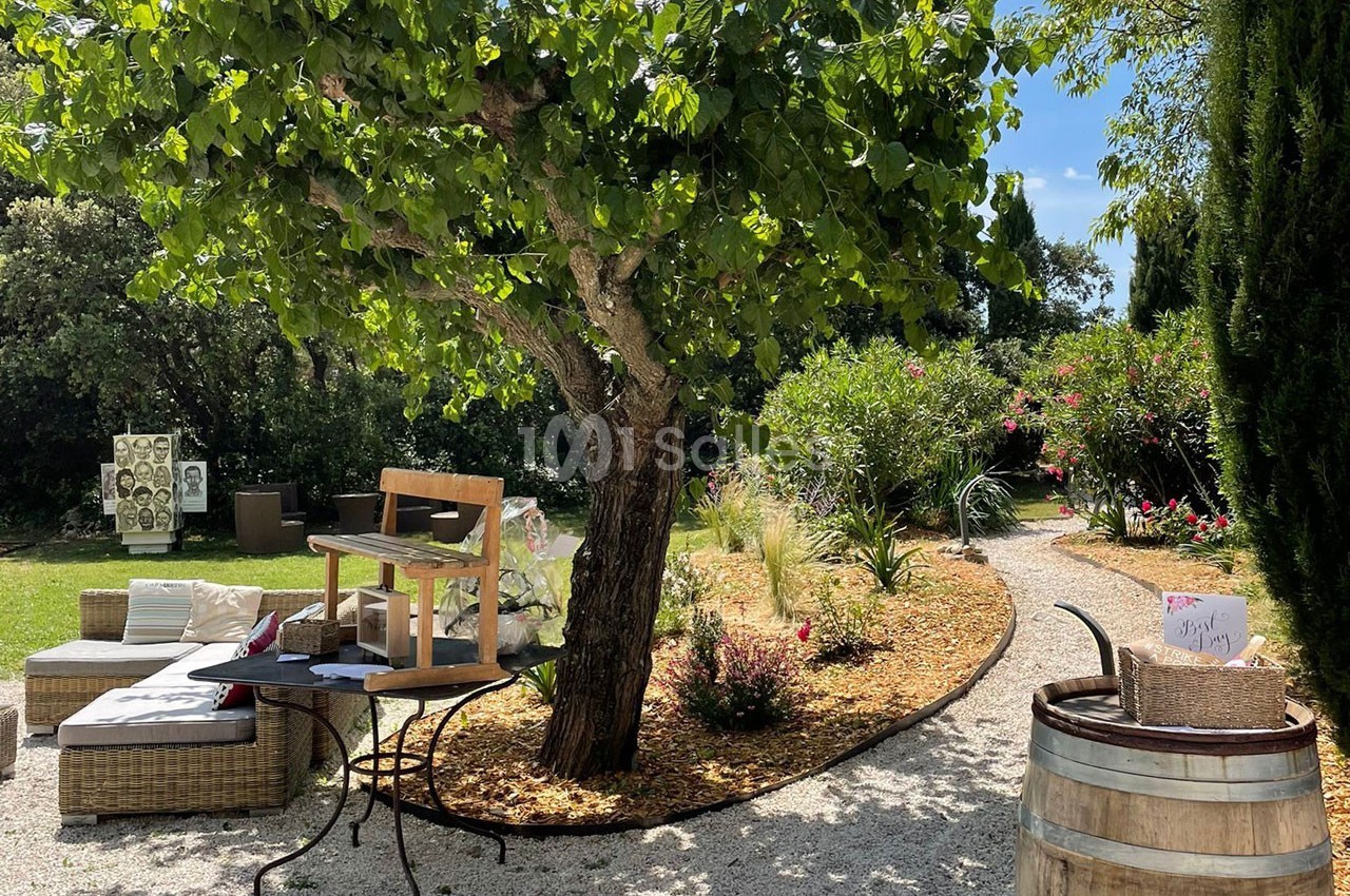 Espace extérieur avec mobilier de jardin sous un arbre, entouré de plantes et d'un chemin gravillonné.