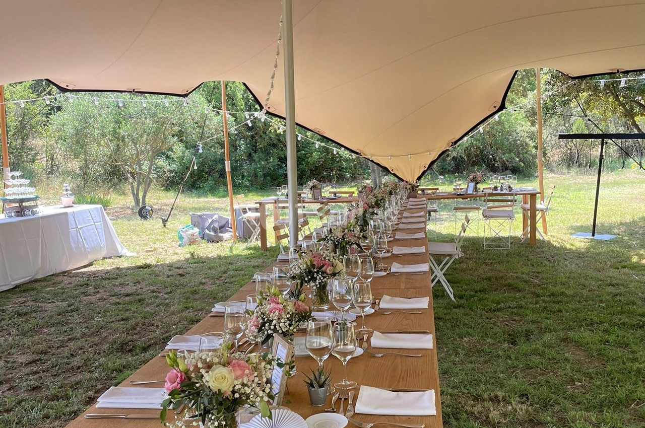 Tables en bois décorées de fleurs et de vaisselle sous une tente blanche dans un cadre extérieur verdoyant.