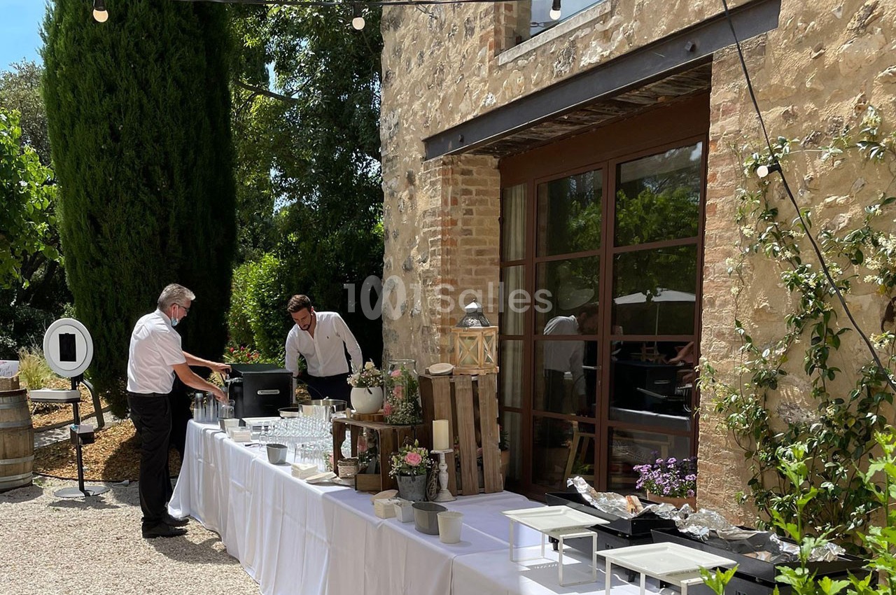 Deux serveurs préparent une table de buffet en extérieur, près d'un bâtiment en pierre entouré de végétation.