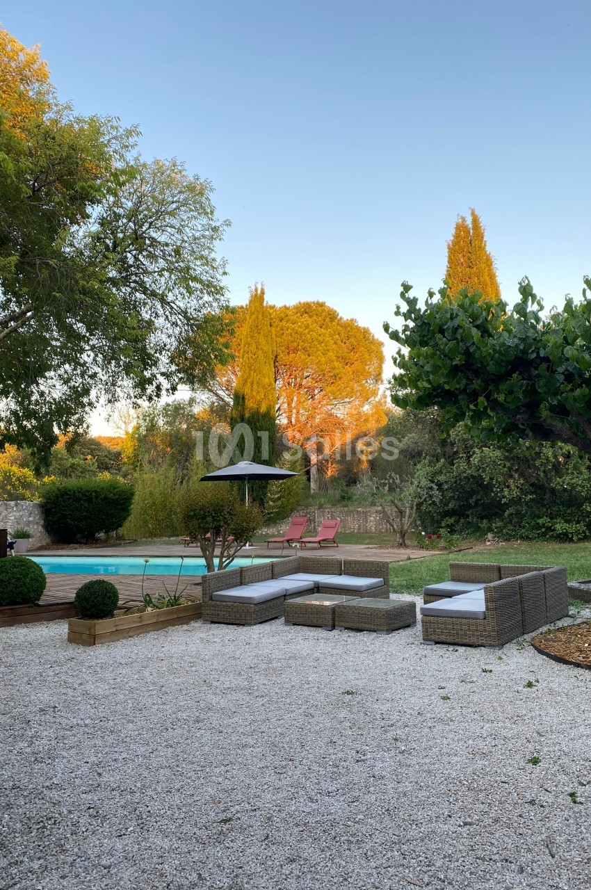 Mobilier de jardin en rotin sur une terrasse en gravier, avec piscine, arbres et chaises longues en arrière-plan.