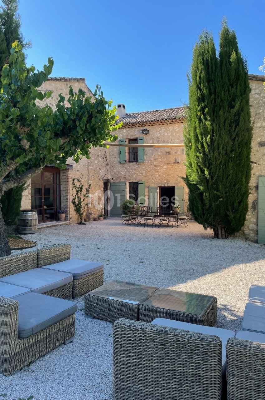 Cour en gravier avec mobilier en rotin, table et chaises en fer forgé, entourée de bâtiments en pierre et de cyprès.
