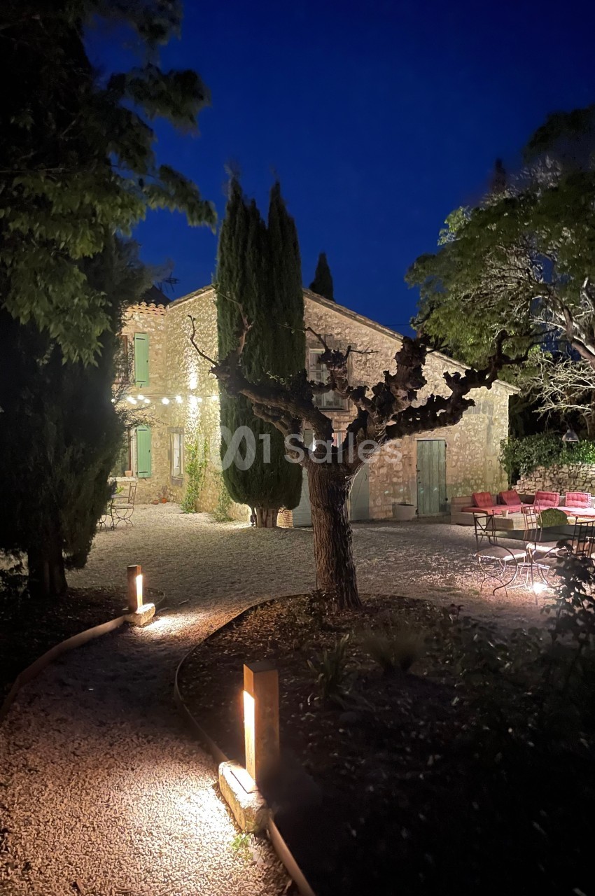 Cour d'une maison en pierre éclairée la nuit, avec arbres, mobilier de jardin et chemin bordé de lampes.