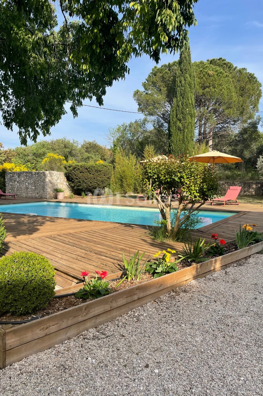 Piscine extérieure entourée de végétation, terrasse en bois, transats rouges et parasol jaune sous un ciel dégagé.