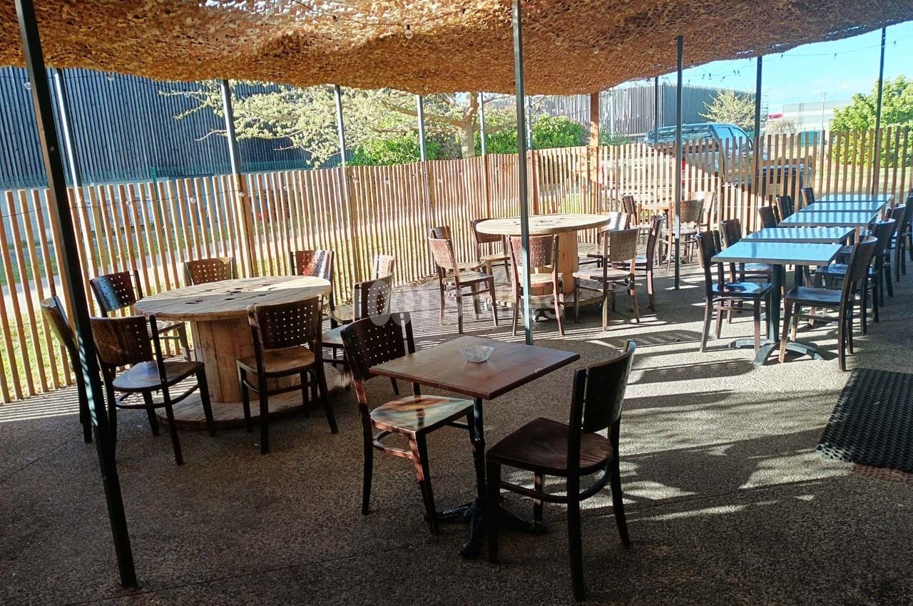 Terrasse extérieure ombragée avec des tables et chaises en bois, entourée d'une clôture en bois clair.