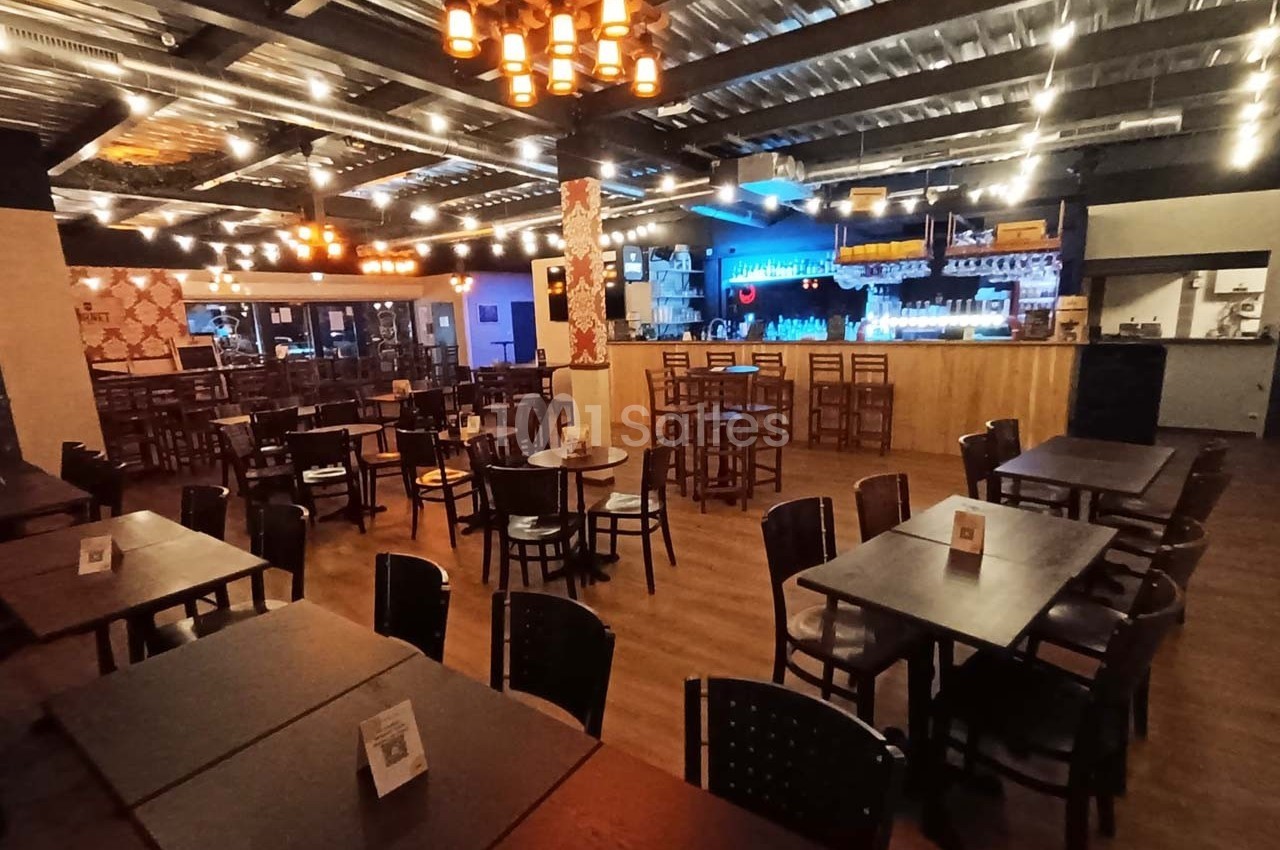 Salle de restaurant spacieuse avec tables en bois, éclairage chaleureux et bar illuminé en arrière-plan.