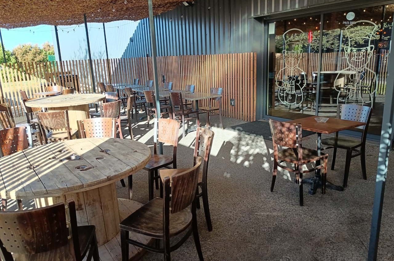 Terrasse extérieure avec tables en bois rondes et carrées, chaises en métal, ombragée par une pergola et clôturée en bois.