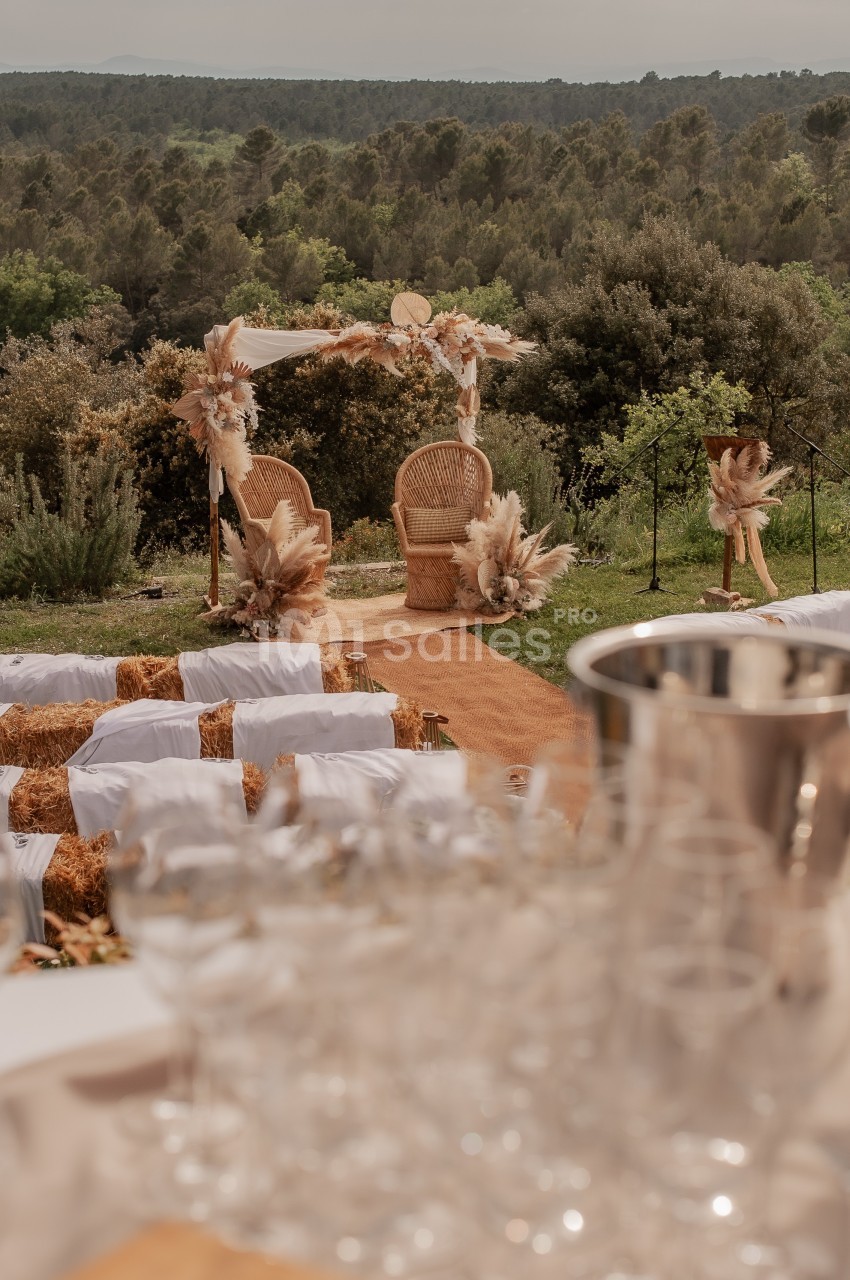 Chaises décorées sous une arche florale dans un cadre naturel, avec des rangées de bottes de paille en arrière-plan.