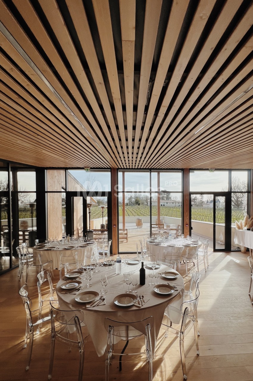 Salle lumineuse avec tables rondes dressées, chaises transparentes et grandes baies vitrées donnant sur un extérieur…