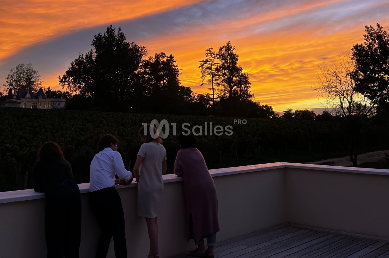 Quatre personnes regardent un coucher de soleil aux teintes orange et violet depuis une terrasse surplombant des vignes.