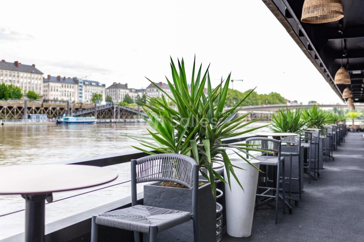 Terrasse avec tables, chaises et plantes, donnant sur une rivière bordée de bâtiments et d'un pont au loin.