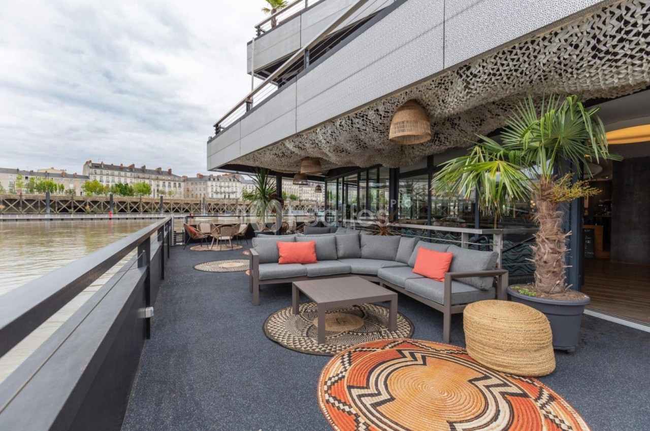 Terrasse aménagée avec canapés, tables basses, plantes et vue sur un fleuve bordé de bâtiments urbains.