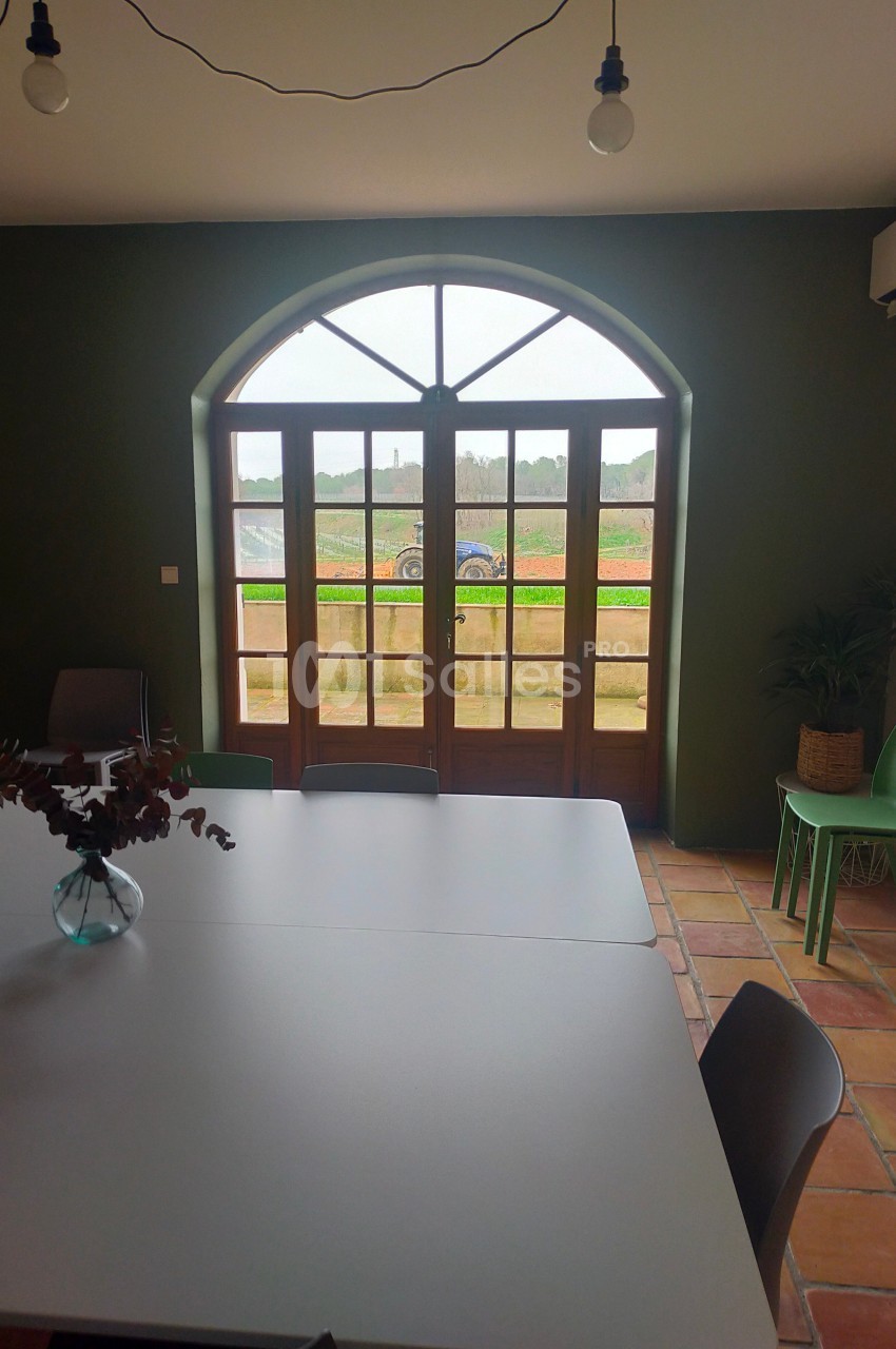 Grande baie vitrée en arche donnant sur un paysage champêtre, vue depuis une salle avec table et chaises.