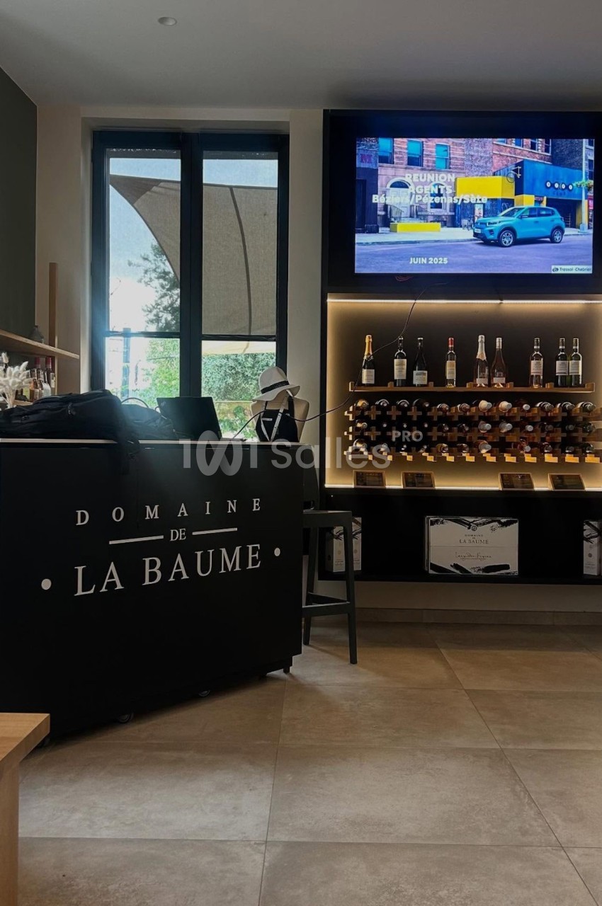 Comptoir noir avec inscription ’Domaine de La Baume’, étagères de bouteilles de vin et écran affichant une publicité.