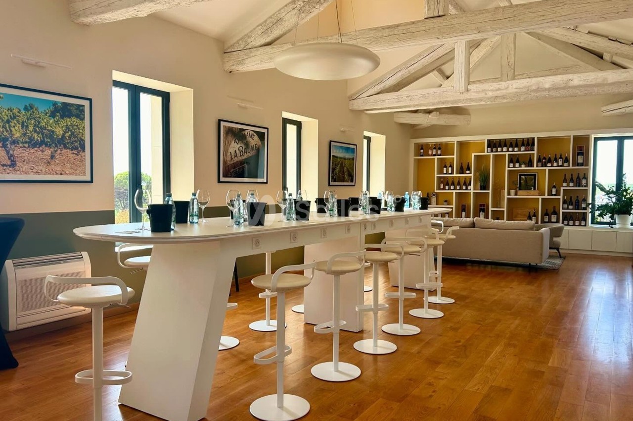 Salle lumineuse avec un long comptoir blanc, des tabourets, des bouteilles exposées et des poutres apparentes au plafond.