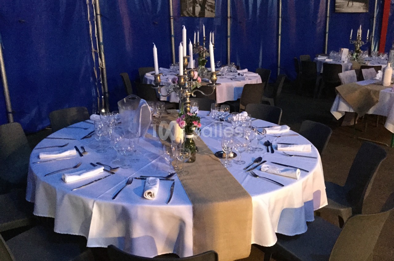 Table ronde dressée avec nappes blanches, couverts, chandeliers et décorations florales, dans une salle aux murs bleus.