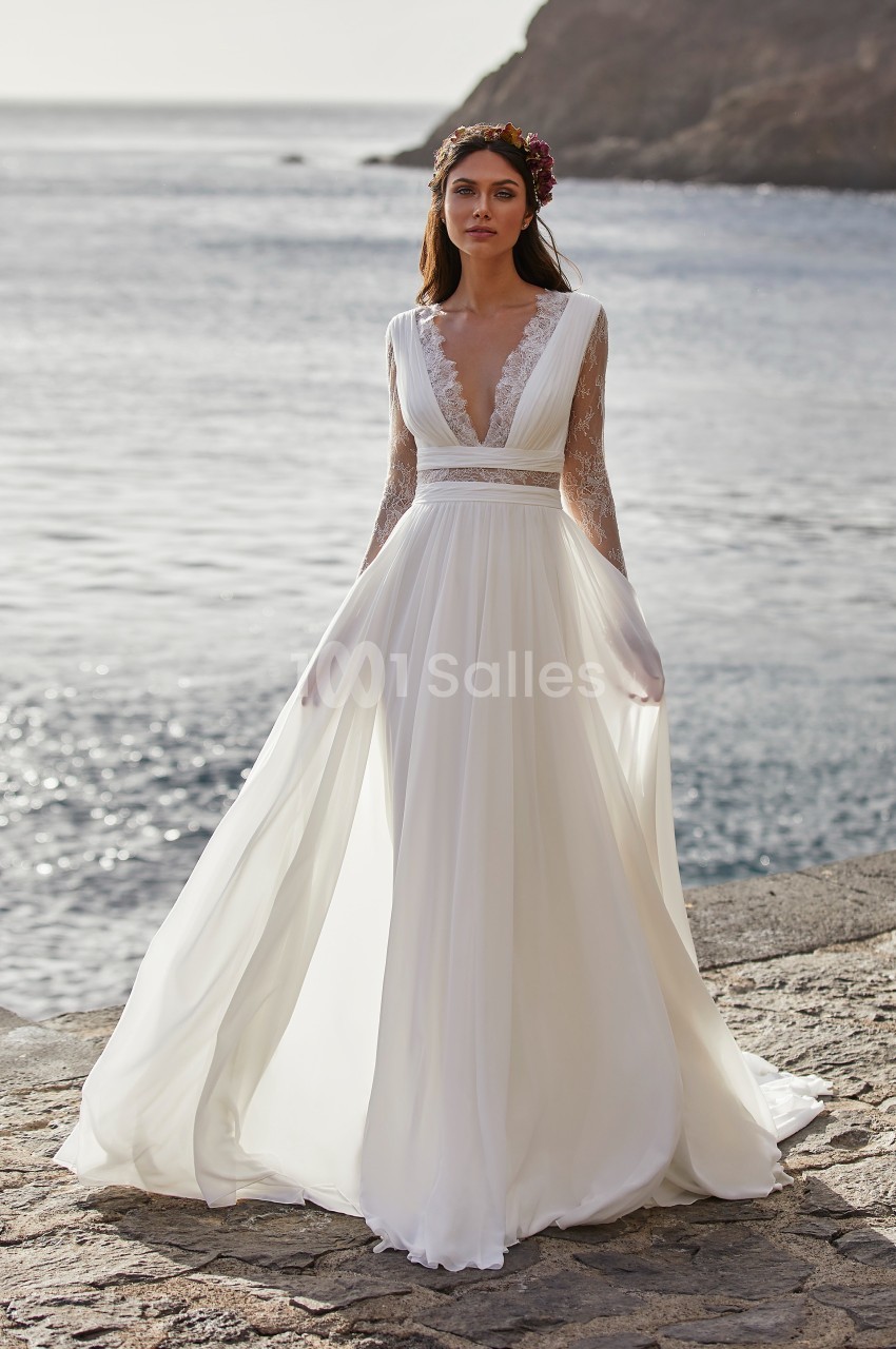 Femme portant une robe de mariée fluide avec dentelle, debout près d'une étendue d'eau et de rochers.