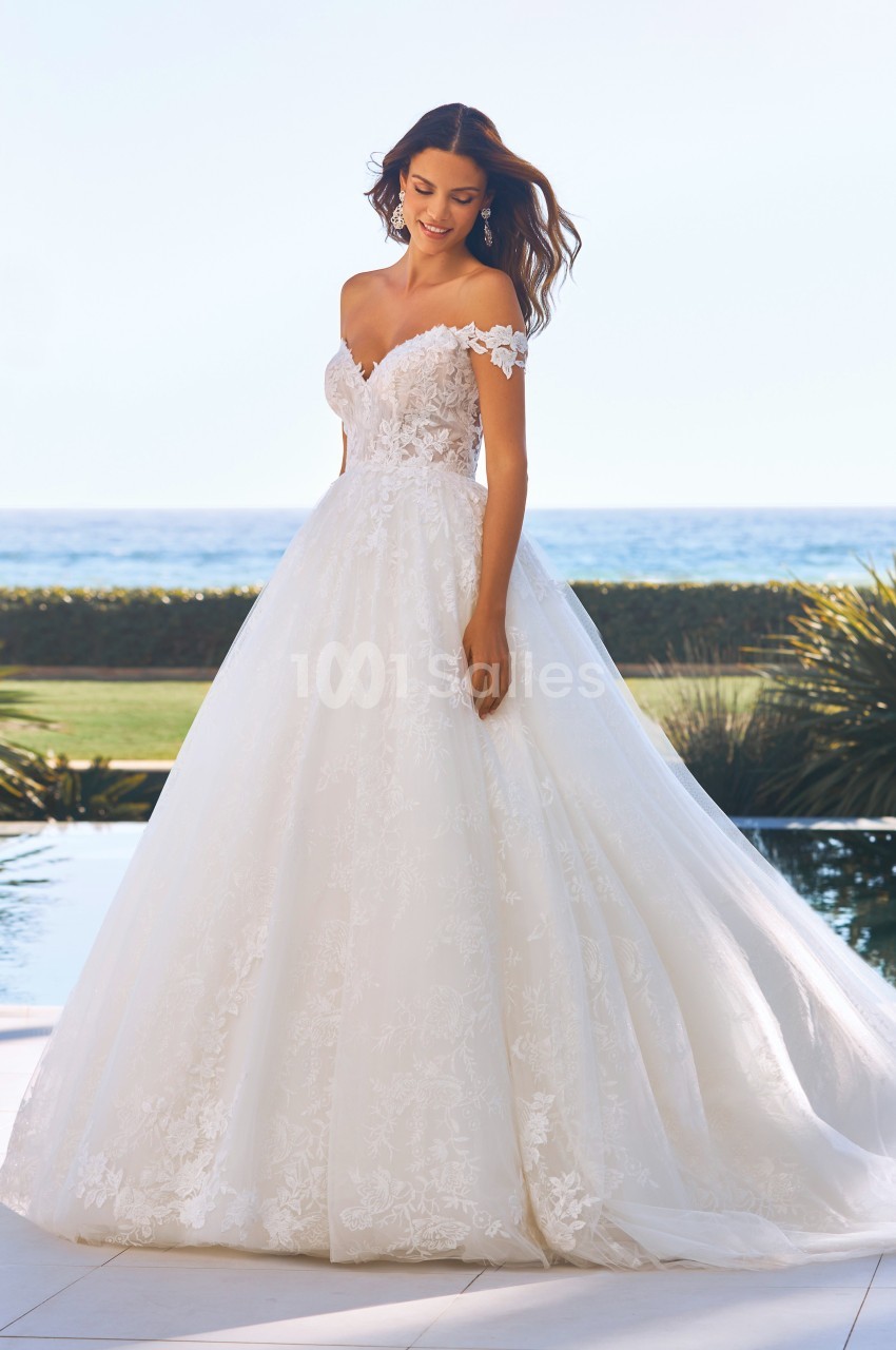 Femme portant une robe de mariée blanche avec dentelle, posant en extérieur près d'une piscine et d'un paysage côtier.