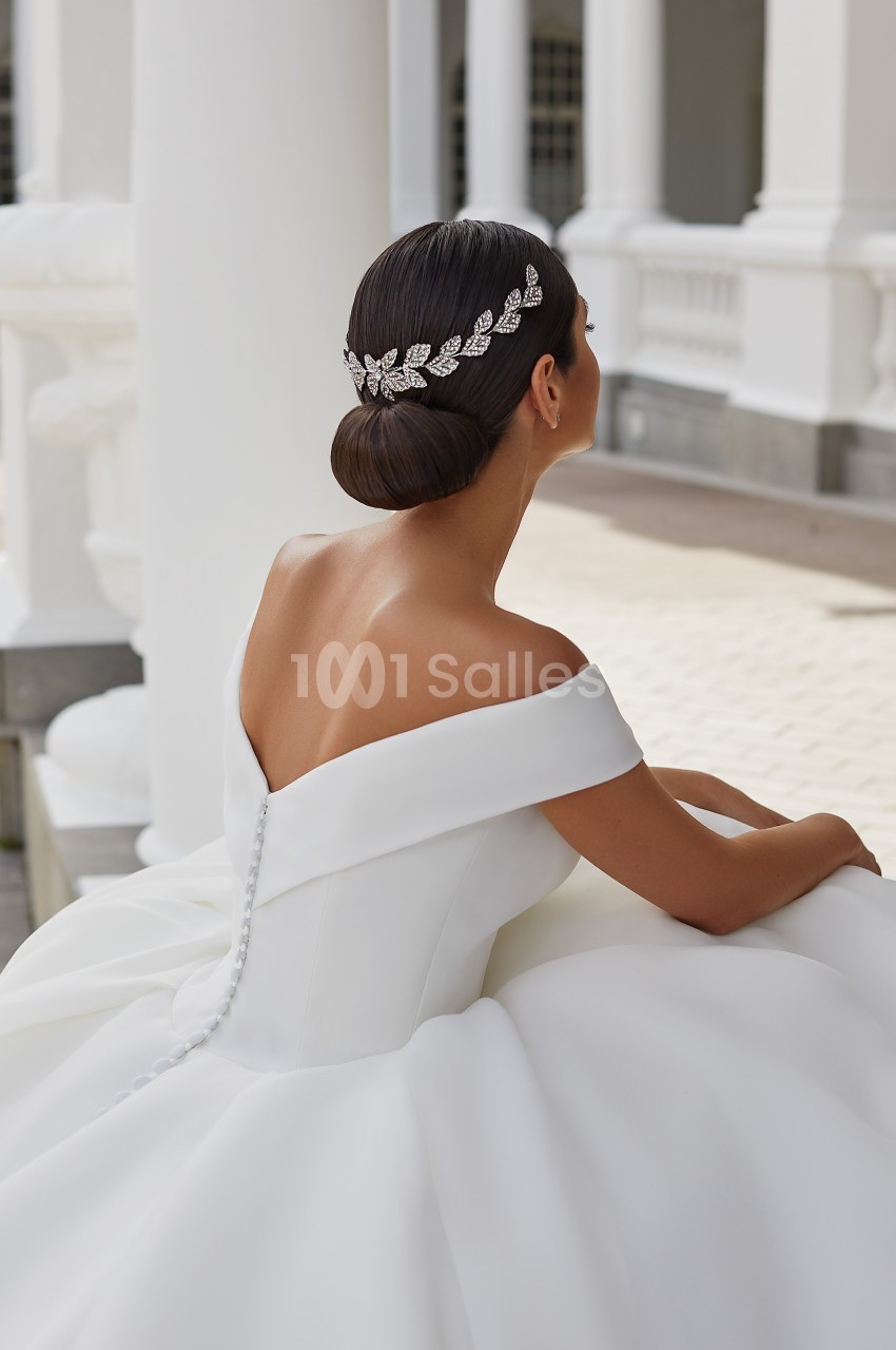 Femme en robe de mariée blanche avec un chignon orné d'un accessoire argenté, assise près d'une architecture classique.