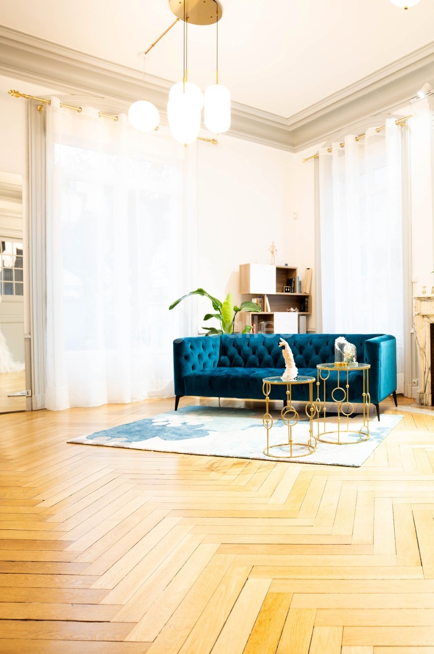 Salon lumineux avec parquet en chevrons, canapé bleu, tables dorées, miroir doré et plantes décoratives.