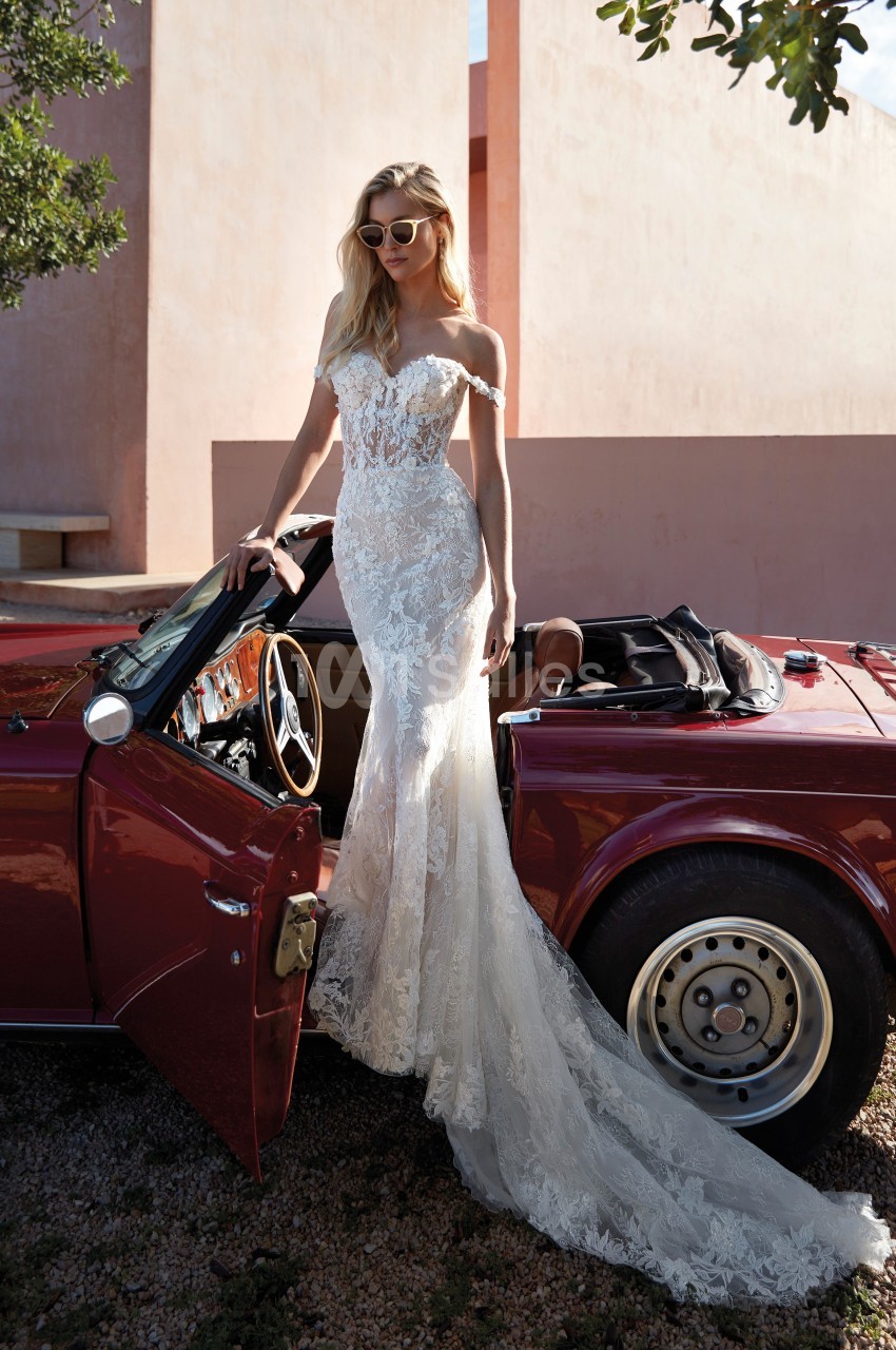 Femme en robe de mariée en dentelle, debout près d'une voiture décapotable rouge dans un décor extérieur ensoleillé.