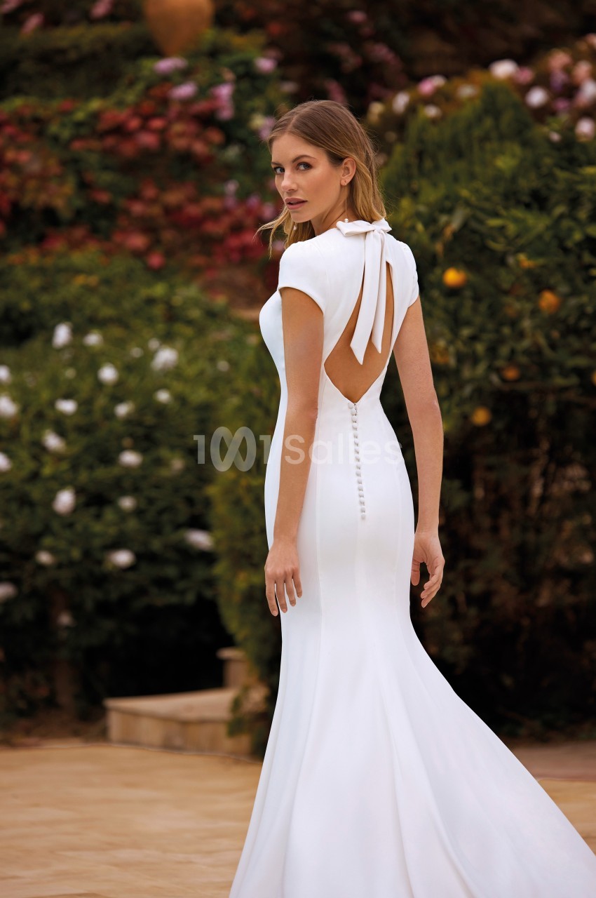 Femme portant une robe de mariée blanche avec un dos ouvert, posant dans un jardin fleuri.