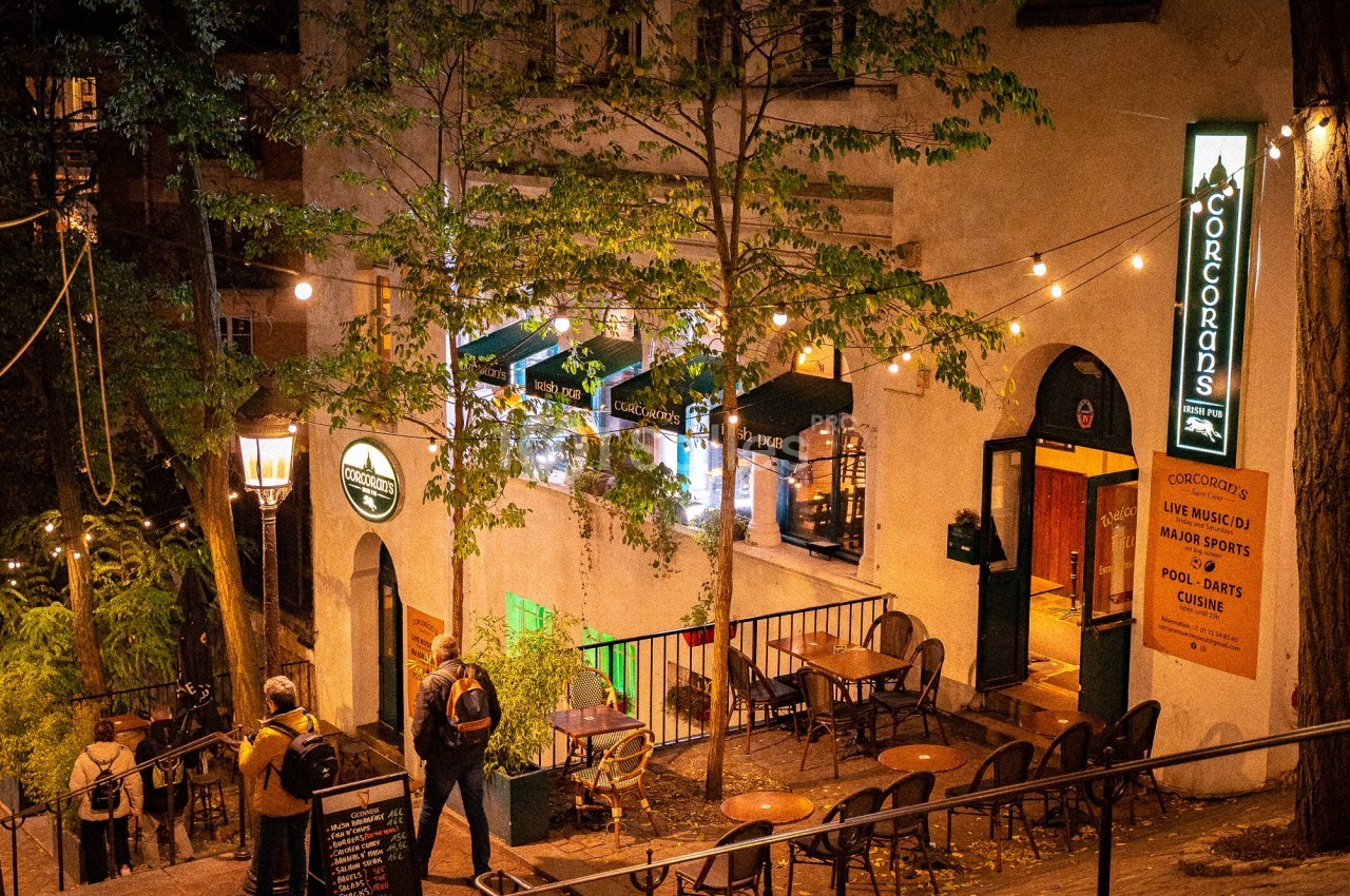 Terrasse extérieure d'un pub éclairé par des guirlandes lumineuses, avec des clients et des arbres environnants.
