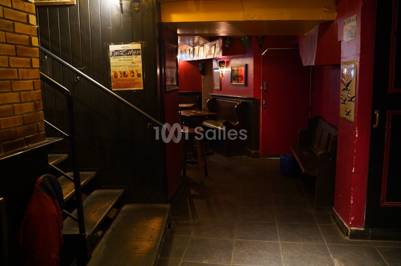 Couloir sombre avec murs rouges et noirs, affiches accrochées, escalier à gauche et mobilier de bar en bois.