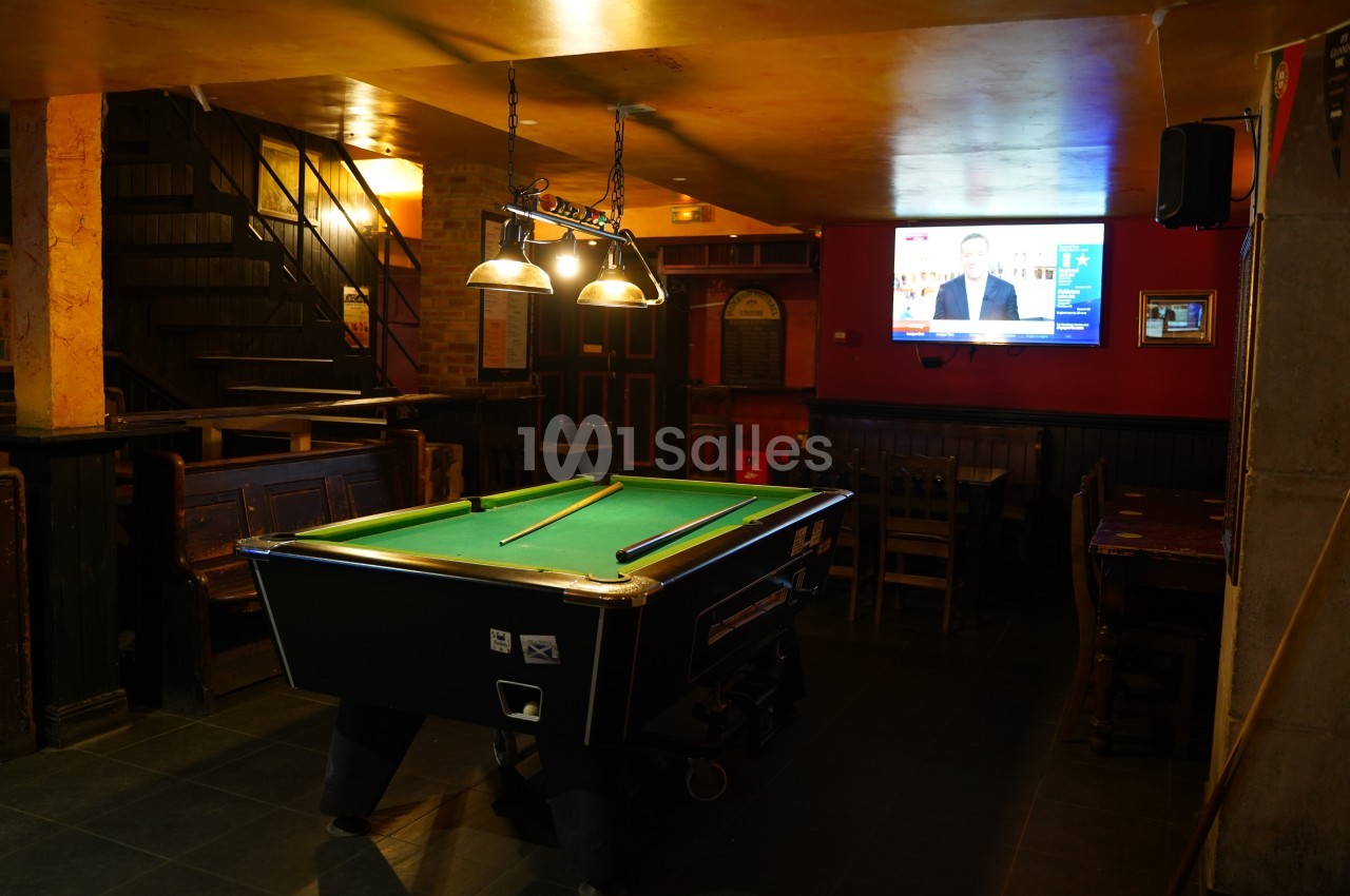 Table de billard éclairée dans un bar à l'ambiance tamisée, avec un écran de télévision allumé en arrière-plan.