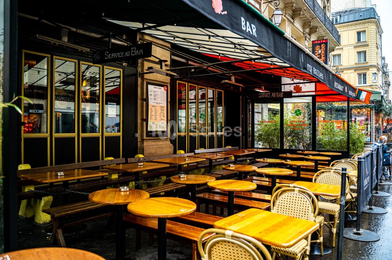 Terrasse de café avec tables et chaises en bois sous un auvent noir, située dans une rue urbaine animée.