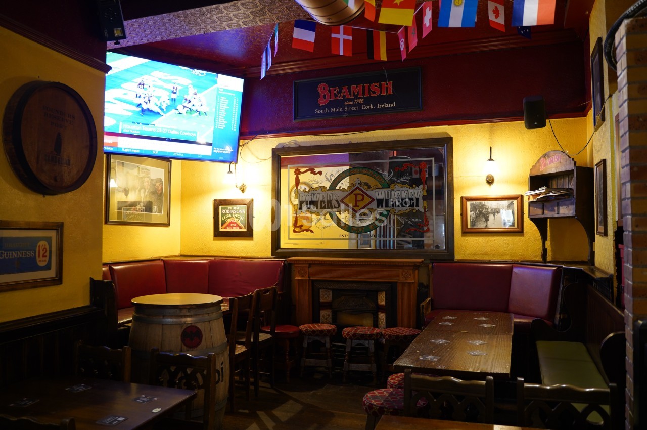 Intérieur d'un pub avec des tables en bois, des banquettes rouges, des drapeaux suspendus et un écran diffusant un match.