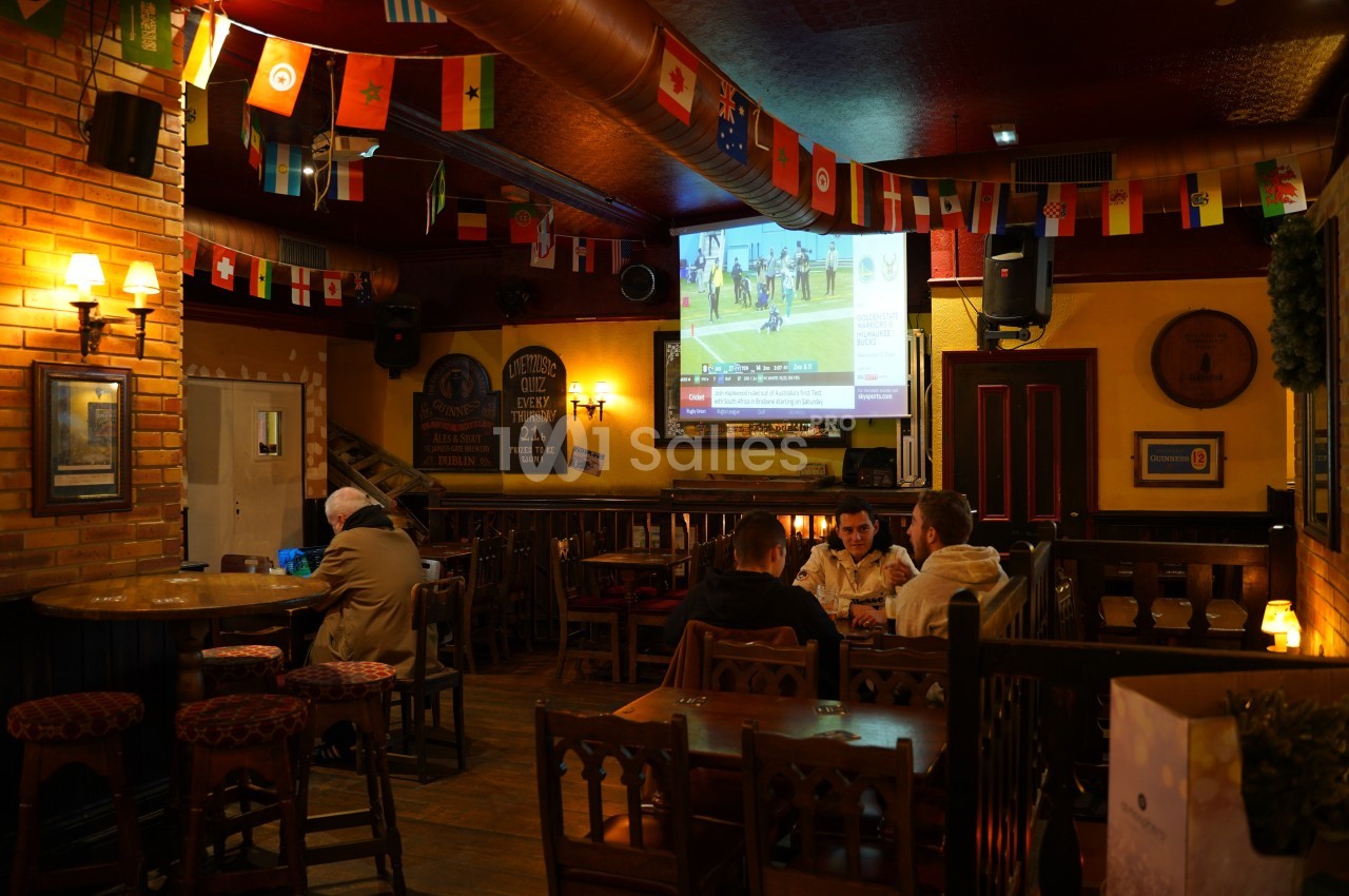Intérieur d'un pub avec des drapeaux suspendus, un écran diffusant un match et des personnes assises autour de tables.