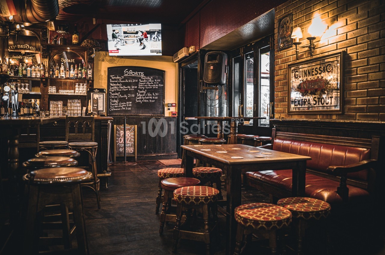 Intérieur d'un pub avec bar, tabourets, tables en bois, éclairage chaleureux et écran diffusant une émission sportive.