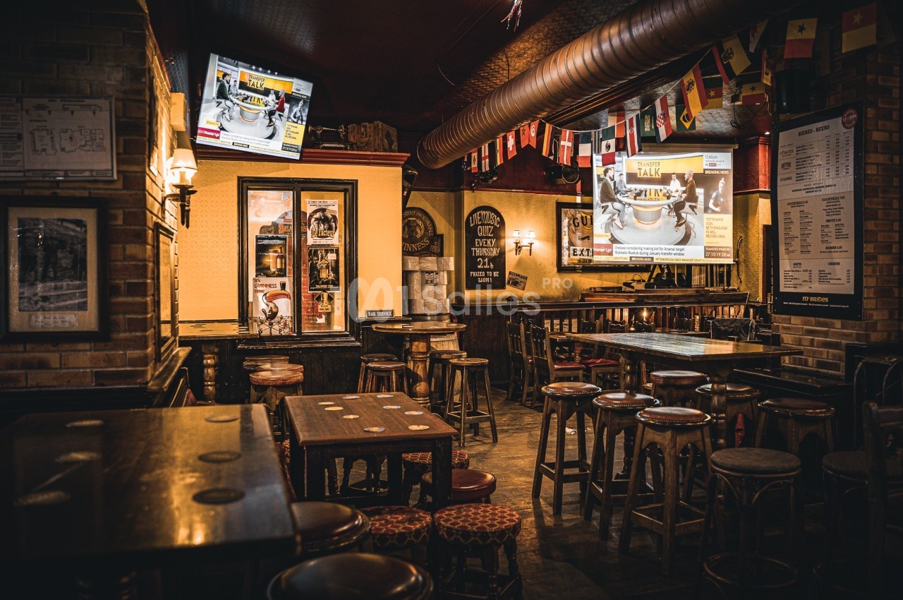 Intérieur d'un bar chaleureux avec tables en bois, tabourets hauts, écrans allumés et décorations de drapeaux.