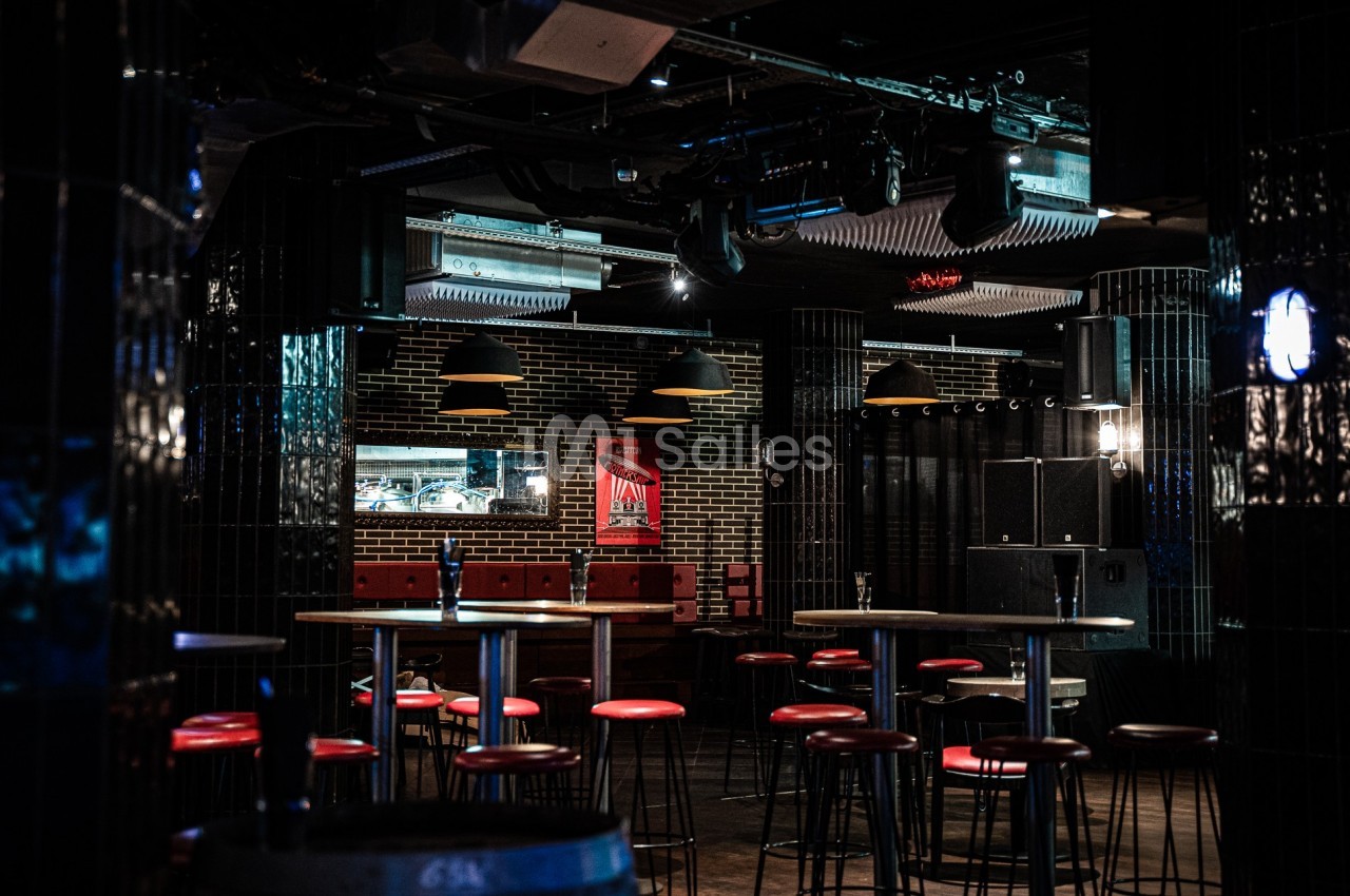 Salle de bar sombre avec tabourets rouges, tables hautes et murs en briques, éclairée par des suspensions noires.