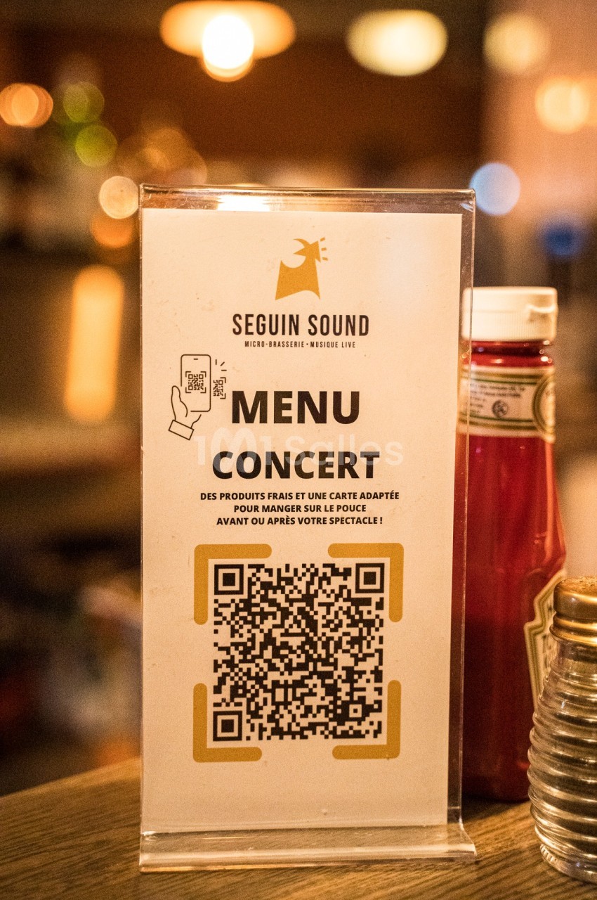 Affiche sur un comptoir présentant un menu concert avec QR code, ketchup et décor flou en arrière-plan.