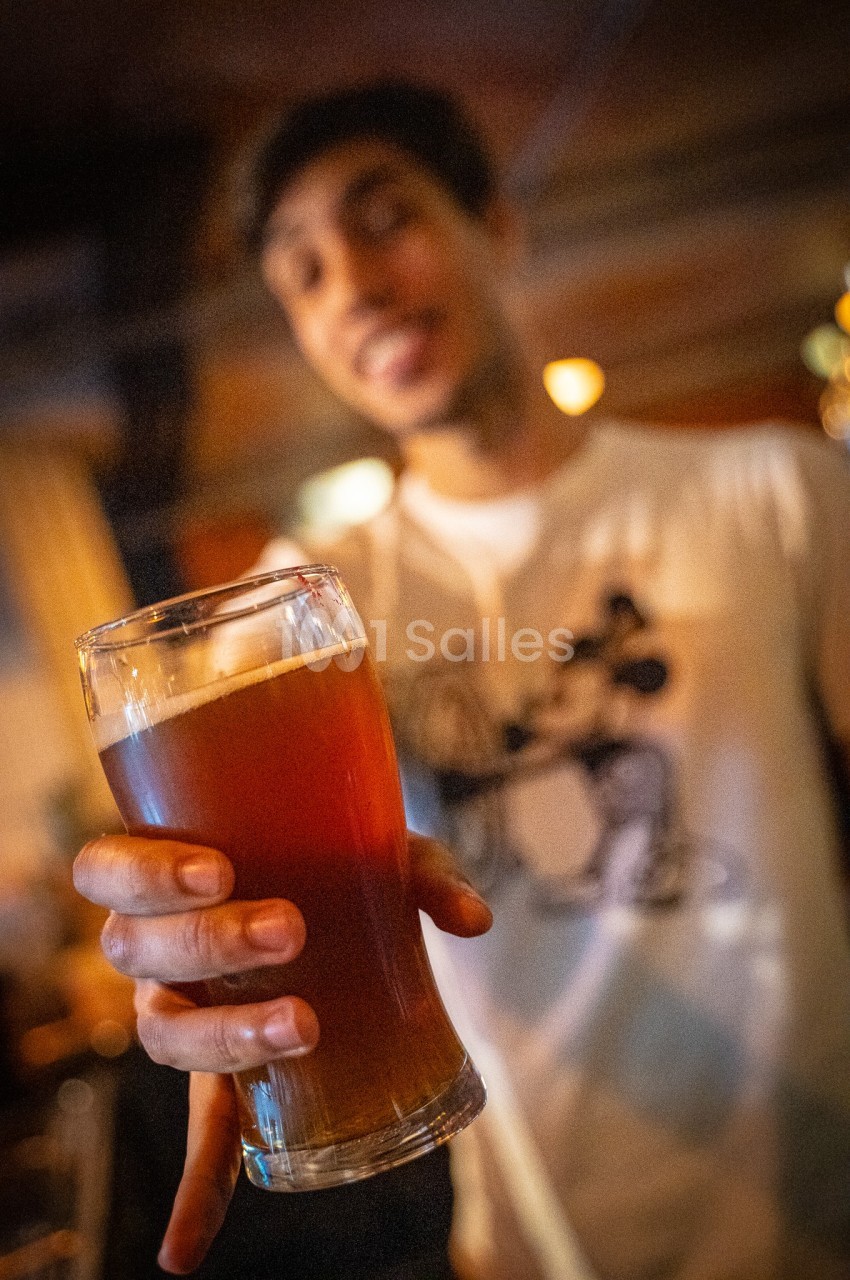 Un homme tend un verre de bière ambrée dans un environnement de bar ou de café.
