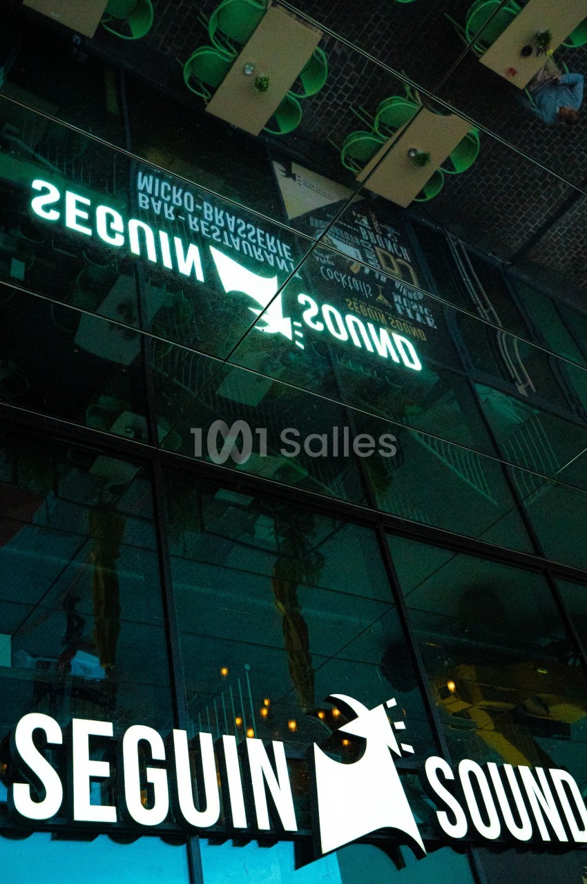 Reflet d'une enseigne lumineuse ’Seguin Sound’ sur une façade vitrée, avec des éléments urbains en arrière-plan.