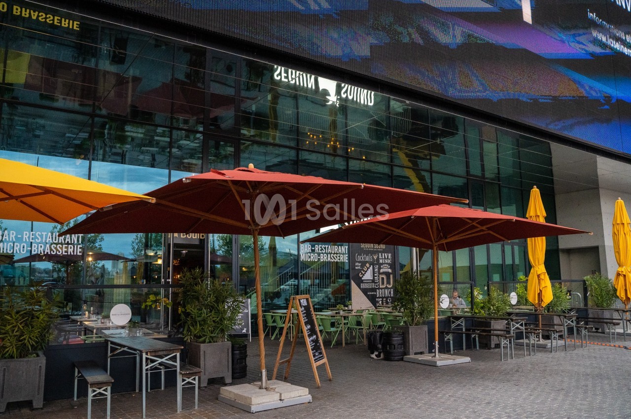 Terrasse d'un restaurant avec parasols rouges et jaunes, entourée de plantes, devant une façade vitrée moderne.