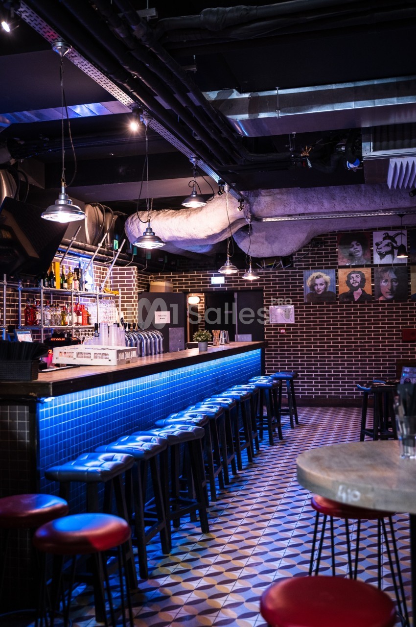Bar intérieur avec comptoir éclairé en bleu, tabourets alignés, murs en briques et décorations murales.