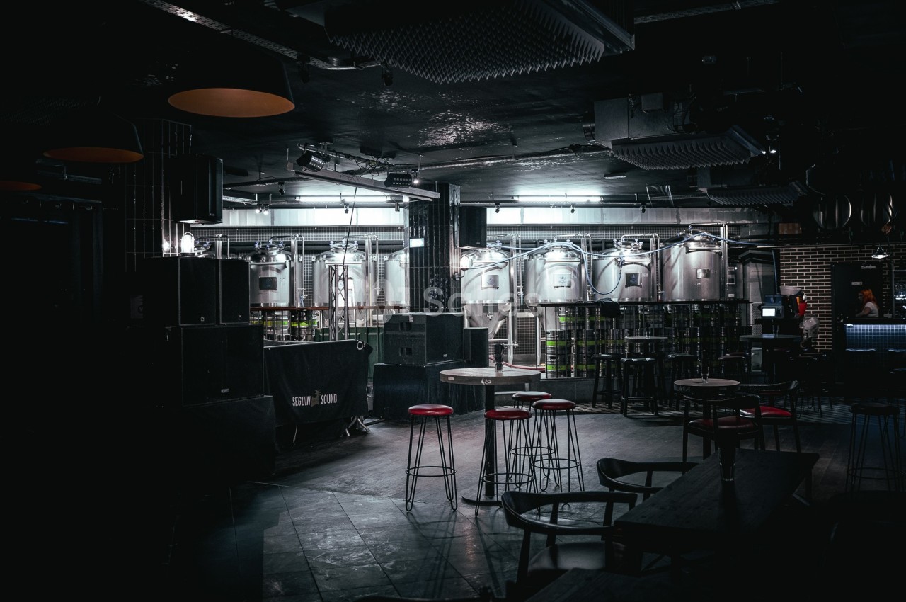 Salle sombre avec des cuves en acier inoxydable, des tabourets rouges et des tables hautes dans un décor industriel.