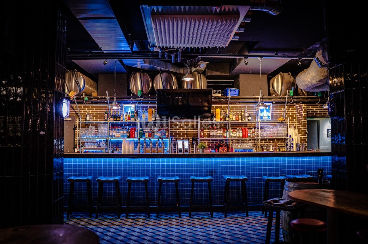 Bar avec comptoir éclairé en bleu, tabourets alignés, étagères remplies de bouteilles et décor industriel en arrière-plan.