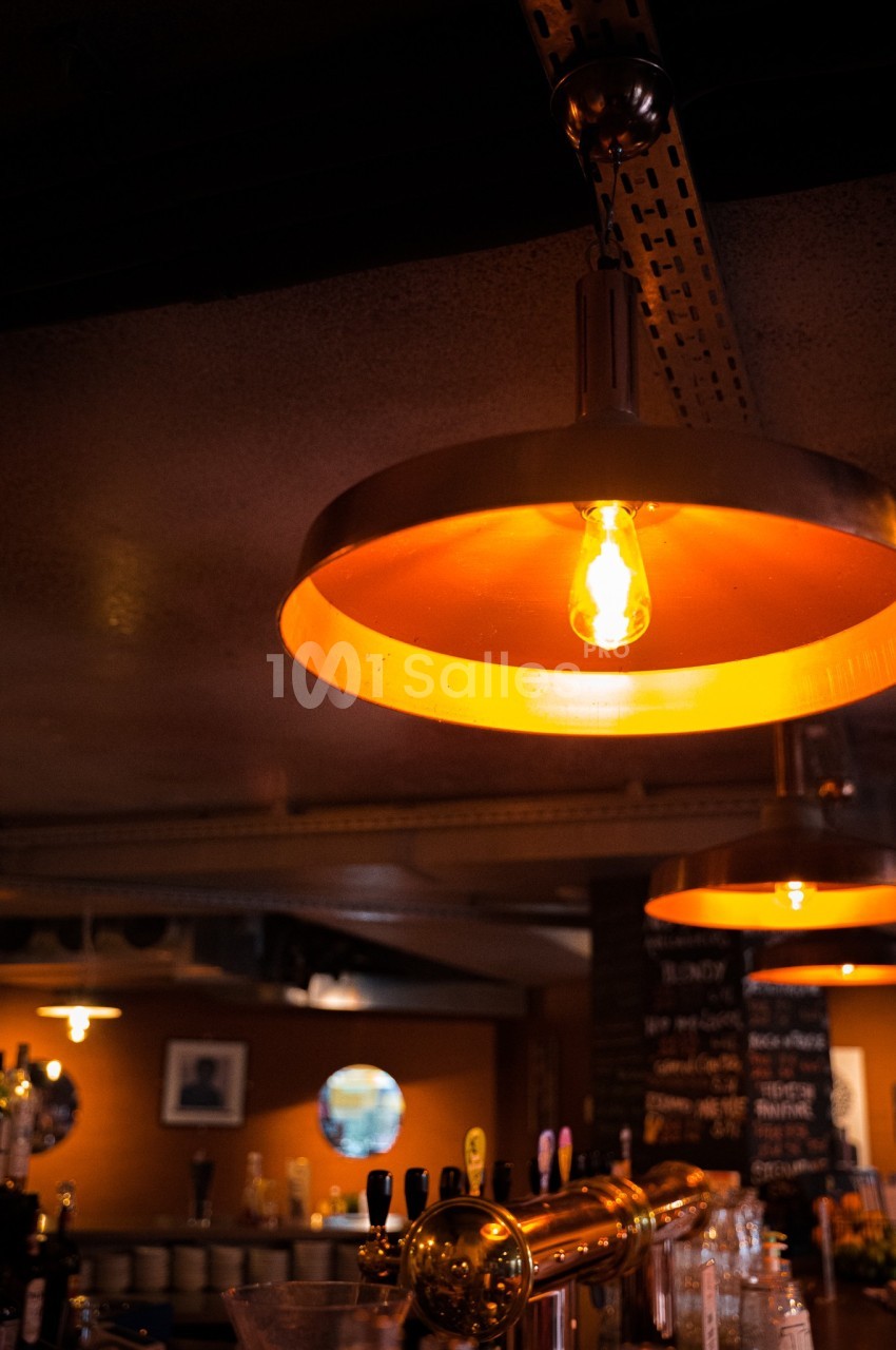 Suspension métallique orange éclairée, suspendue au plafond d'un bar avec un comptoir et des bouteilles en arrière-plan.