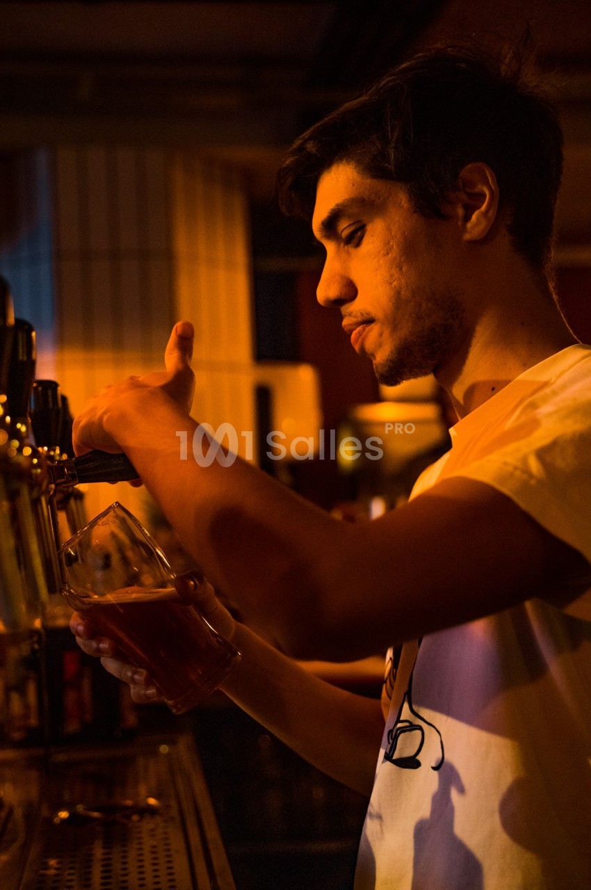 Un barman sert une bière pression dans un verre, éclairé par une lumière chaude dans un bar.