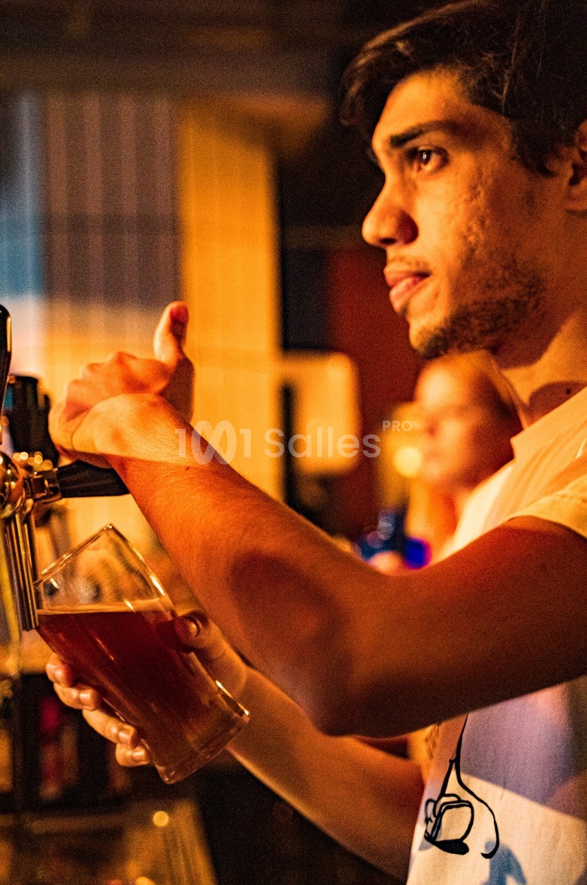 Un homme sert une bière pression dans un verre transparent dans un environnement faiblement éclairé.