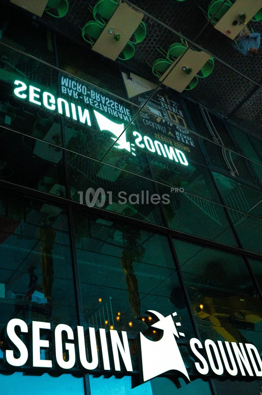 Reflet d'une enseigne lumineuse ’Seguin Sound’ sur une façade vitrée, avec des éléments urbains en arrière-plan.