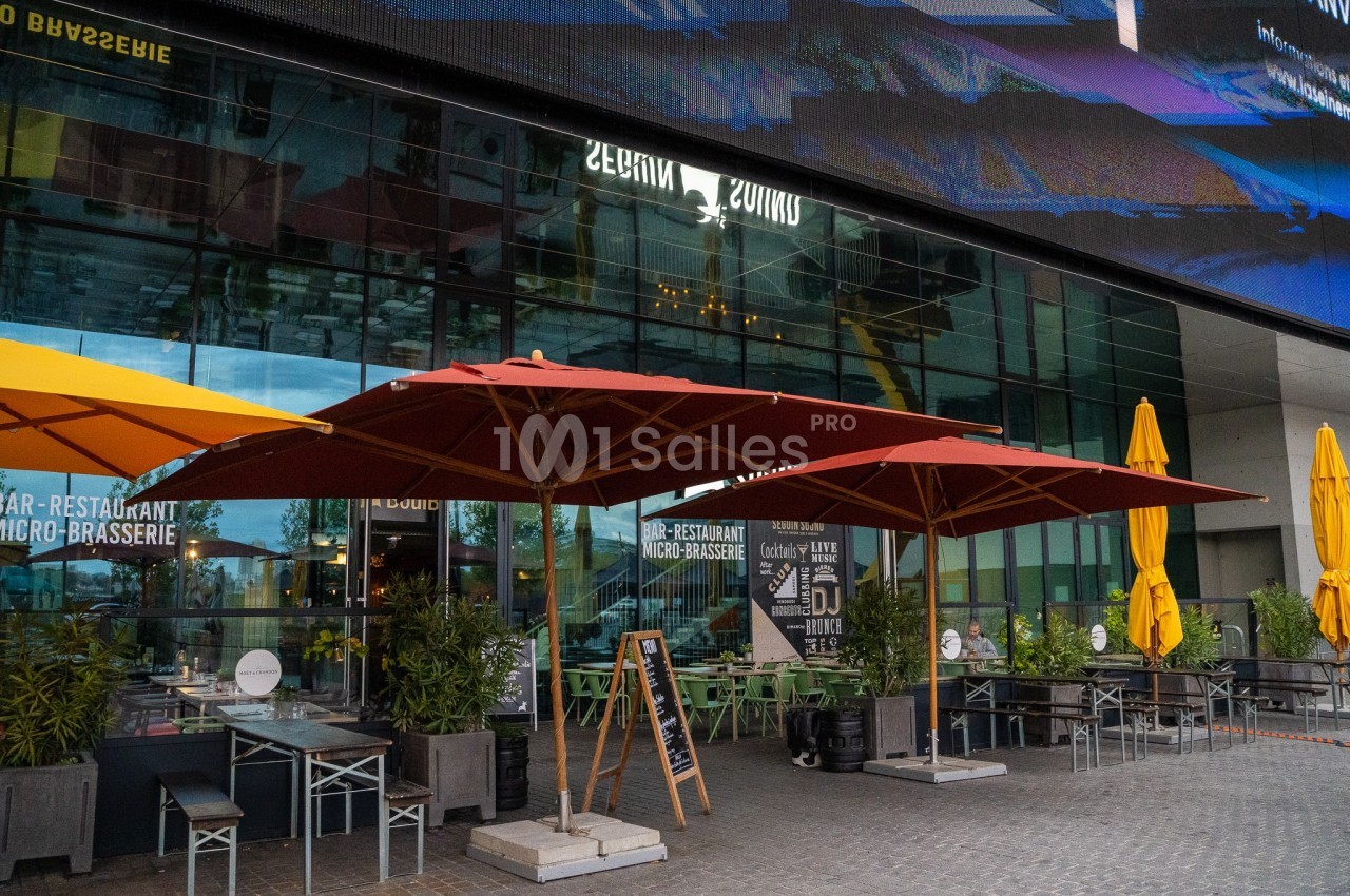 Terrasse d'un restaurant avec parasols rouges et jaunes, entourée de plantes, devant une façade vitrée moderne.