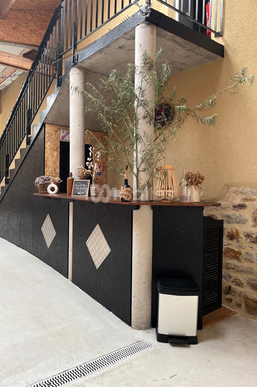 Comptoir en bois et béton décoré de plantes et objets, situé sous un escalier en intérieur avec mur en pierre.